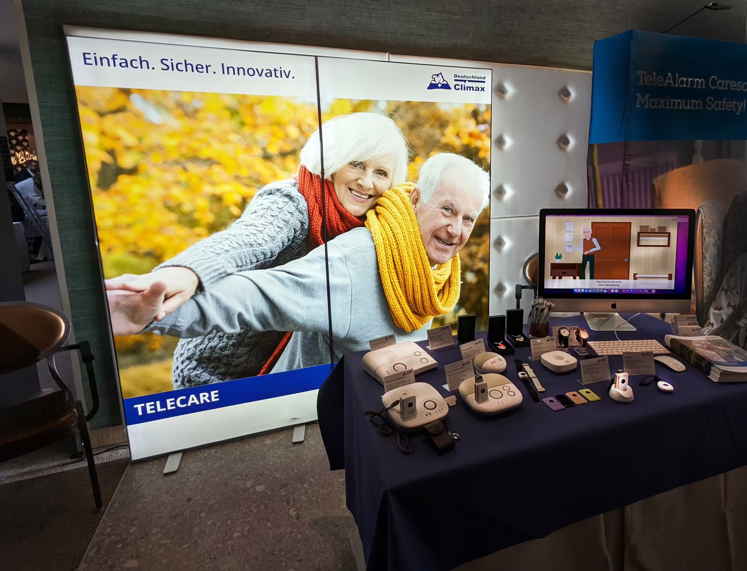 PXL_20230925_065702097_bearbeitet Der Messestand von Climax mit einer Rückwand auf der ein älteres Pärchen zu sehen ist und davor steht ein Tisch mit verschiedenen Hausnotrufgeräten.